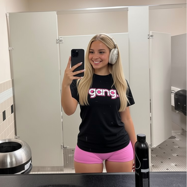 Person taking a mirror selfie in a bathroom wearing a black t-shirt with 'gang' text and pink shorts.