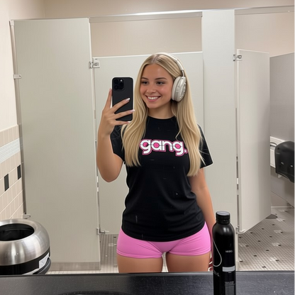 Person taking a mirror selfie in a bathroom wearing a black t-shirt with 'gang' text and pink shorts.