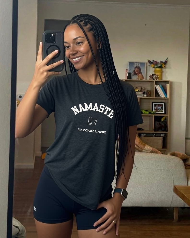 Person wearing a black 'NAMASTE IN YOUR LANE' t-shirt taking a mirror selfie in a living room.